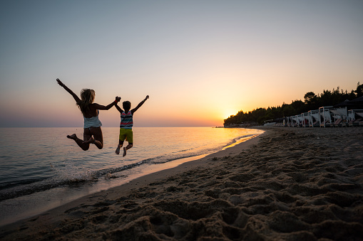 Rear view of playful mother and son holding hands while jumping on the beach in summer day