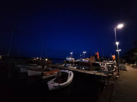 In the idyllic cozy harbor after dark, still light in the horizon from the sinking sun. Various ships docked on the pier like sailboat, motor boat, ferry, commercial ship, yachts and local fisher boats