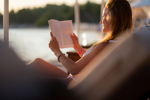 Happy woman reading a book while relaxing on deck chair during summer day on the beach