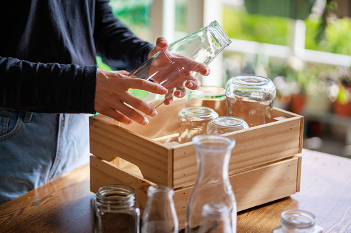 Cropped image of an Asian woman's hands placing empty glass jars into a wooden box, ready for recycling, showcasing a sustainable lifestyle.