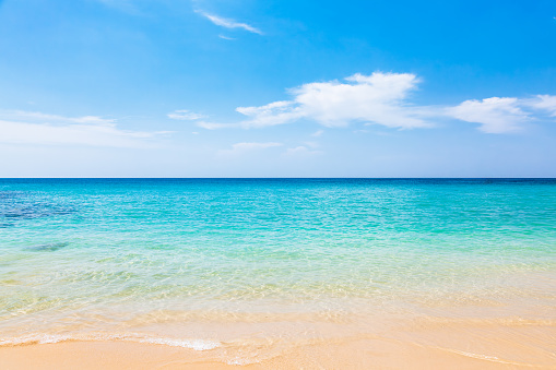 A tropical beach with blue and green water and white sand. The water is a gradient of blue and green, with the horizon in the background. The sand is a light beige color and is smooth and clean. The sky is a bright blue with a few wispy clouds. The overall mood of the image is peaceful and serene.