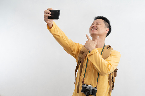 Smiling attractive young tourist with backpack walking and taking selfie mobile phone on white background. Passenger traveling on weekends. Air flight journey concept