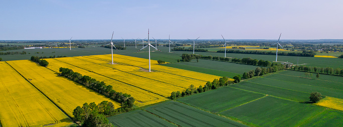 Aerial view Wind turbine on grassy yellow farm canola field against cloudy blue sky in rural area. Offshore windmill park with clouds in farmland Poland Europe. Wind power plant generating electricity. Renewable green clean energy. Sustainable living