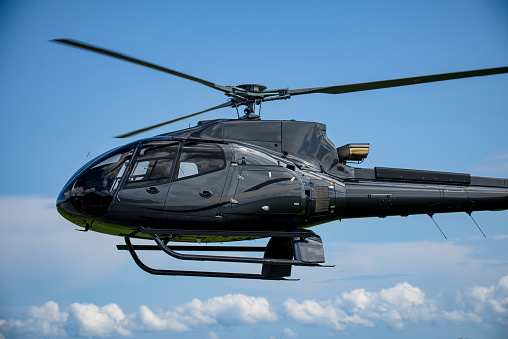 Dark metallic grey helicopter in flight, set against a blue sky with clouds in the lower third. Selective focus used to soften details in the sky,