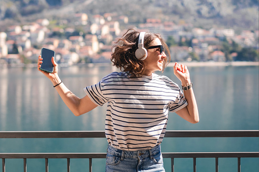 Young happy woman listening to music using headphones while standing on the balcony with  lake and mountain views