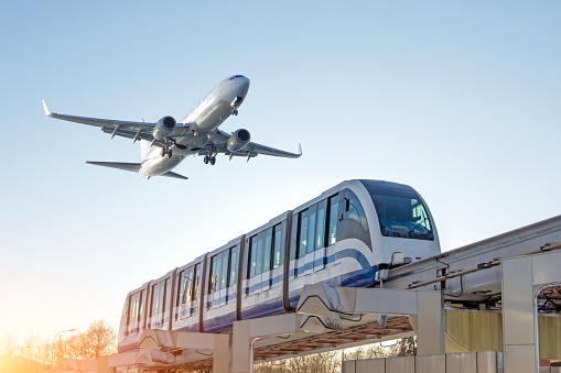 View railway track and suburban electric monorail train rushing to . Passenger plane flying in sky, landing at airport. Concept of modern infrastructure transport travel.