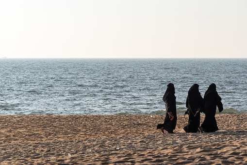 Ullal, India - January 19 2023: Three muslim women in traditional clothing walking on a beach.