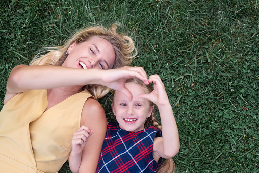 Smiling mother and little daughter lie on green grass on sunny summer day young woman and girl make heart symbol with hands with happy expressions on faces upper view