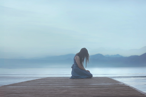 lonely woman kneeling on a pier on the sea lets herself be carried away by emotions in a blue atmosphere