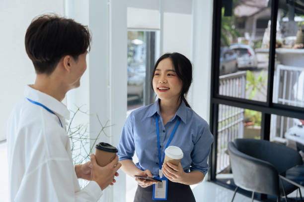 オフィスの窓際でおしゃべりをしたり、話したり、コーヒーを飲んだりする2人の若いアジア人ビジネスウーマン - 女性　会話 ストックフォトと画像