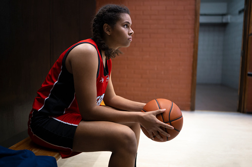 Thoughtful young woman looking away while sitting with basketball on bench in sports hall.