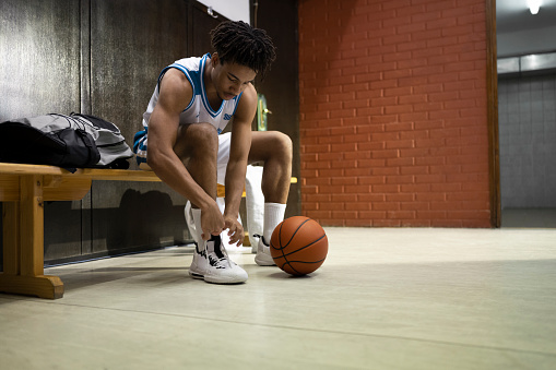 Young basketball player tying shoelaces while sitting on bench in sports hall.
