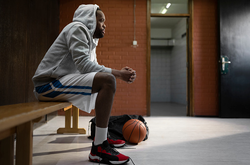 Thoughtful basketball player wearing hooded top looking away while sitting on bench in sports hall.