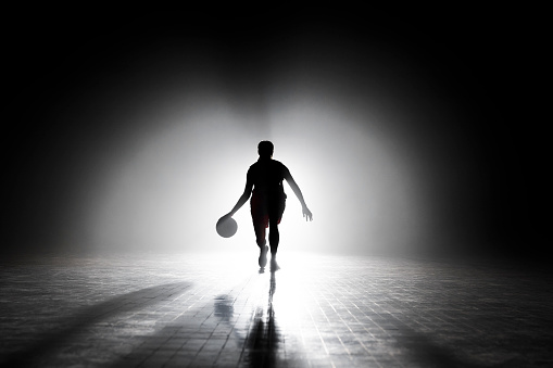 Silhouette of female basketball player dribbling while playing basketball on sports court during practice.