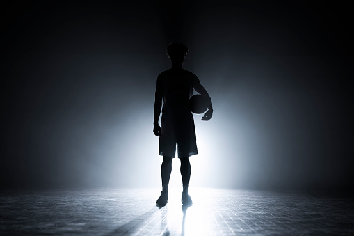 Silhouette of male basketball player standing with basketball on sports court during practice.