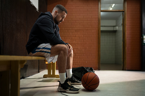 Thoughtful young basketball player looking at basketball kept of floor while sitting on bench in sports hall.