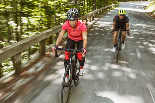 Portrait of a mid-adult woman and man in cycling gear. Helmet, sunglases and cycling clothes. Riding through forest on a mountain road. Going down with great speed.
