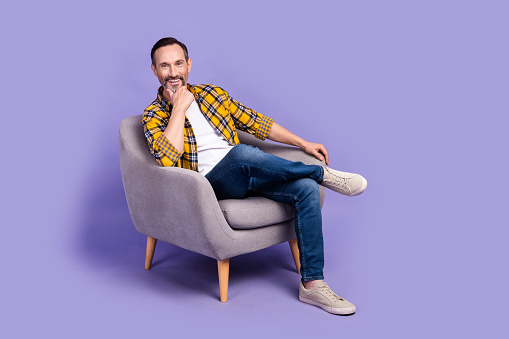 Photo of positive cheerful man wear trendy clothes relax rest chair hand touch chin enjoy comfort isolated on purple color background.