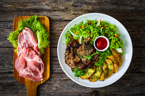 Roast pork neck, fried potato slices and fresh vegetable salad on wooden table