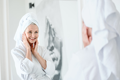 Great result. Focus on mirror reflection of smiling mid age woman in bathrobe touching face, enjoying soft, fresh, healthy look of skin. Beauty care, skincare procedure effect