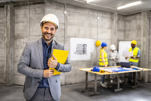 Portrait of an experienced architect with hard hat holding project plan while civil engineers brainstorming in background.