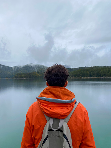 A mobile shot of a mid adult man wearing a waterproof and backpack exploring lake Eibsee while on holiday in Garmisch-Partenkirchen, Germany. He is standing near the water's edge while admiring it with his back to the camera.
