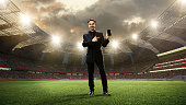 Business in a suit standing on 3d sports arena with flashlights and pointing on mobile phone screen. Betting before game