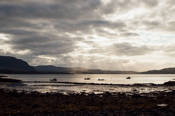 A wide high-contrast shot of a sea loch shoreline with sunbeams illuminating the water in Torridon, Scotland. There are boats in the water and distant hills, with a dramatic cloudy sky.
