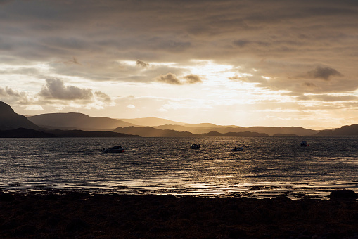 A wide shot of a high-contrast sunset over the shoreline of a sea loch in Torridon, Scotland. There are boats in the water and hills in the distance which are illuminated by the sunlight coming through the clouds.