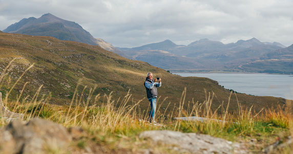 A low-angle wide shot of a senior male adult standing on a hill, taking a photograph of the view. In the distance are rolling hills and mountains, with a sea loch visible to the right of the frame. There are patches of sunlight on the hill in Torridon, Scotland.