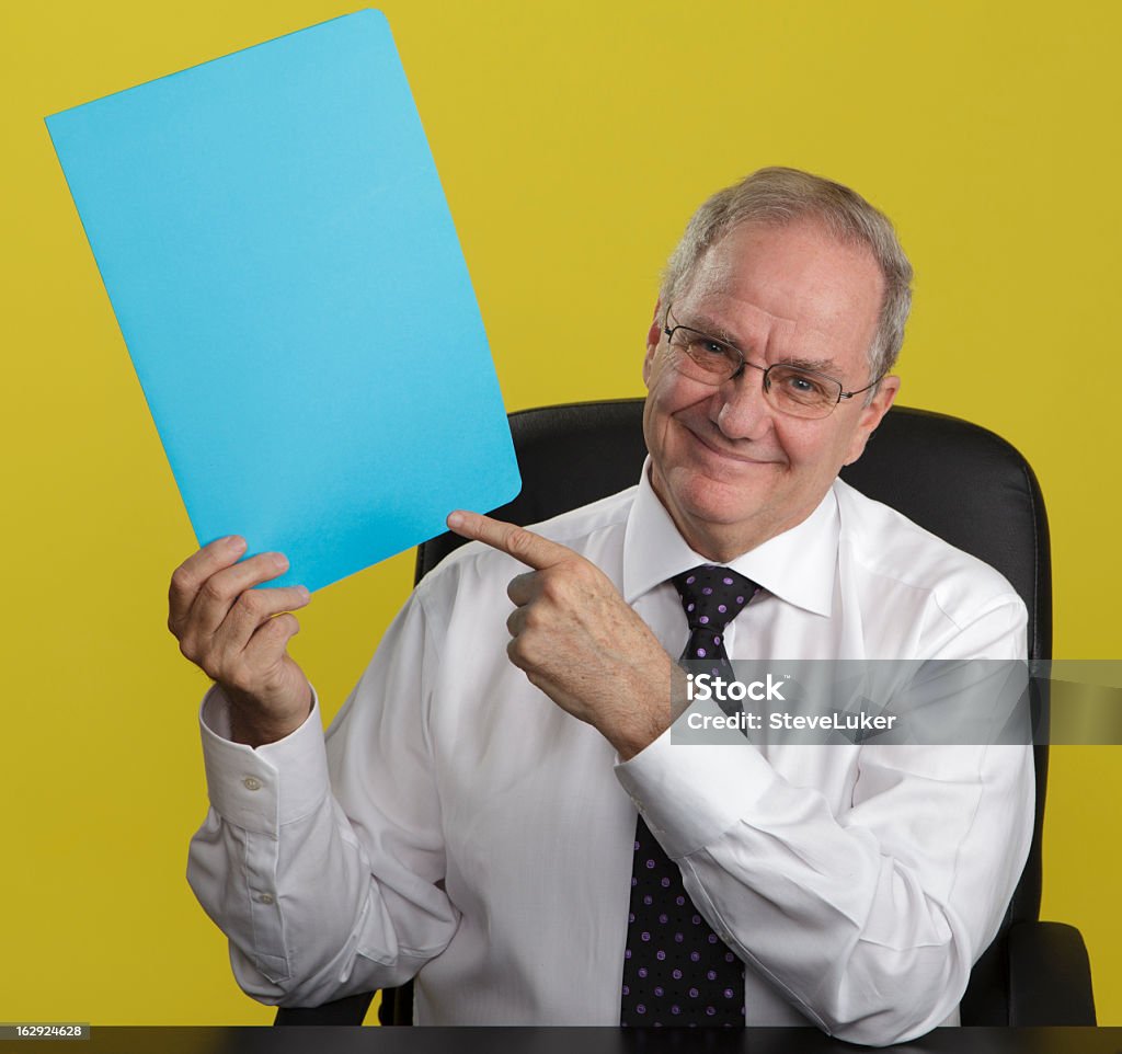 Smiling Businessman Pointing at Blank Sign Smiling businessman sitting at his office desk pointing at a blank blue sign. Men Stock Photo