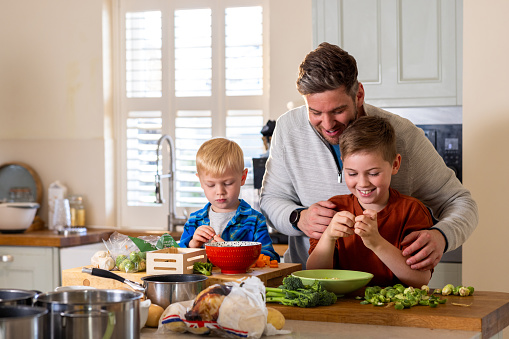 A waist-up shot of a father helping his children prepare dinner together. They are all smiling and talking together around a kitchen island. The kitchen island has lots of fresh vegetables and cooking pans on top of it.