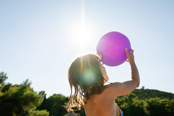 mädchen im grundschulalter, das einen lila ball gegen die sonne hält - sun sky child balloon stock-fotos und bilder