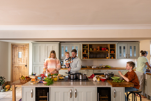 Full shot of a multigenerational family with two children preparing dinner together on a kitchen island in their home. They are all smiling and concentrating on what they are doing. The kitchen island has lots of fresh vegetables on top of it.