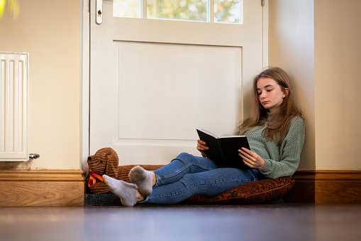 Full shot of a female teenager sitting in a doorway in her home. She is sitting on a cushion, concentrating on a book she is reading.