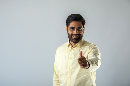 Portrait of smiling handsome young man with beard and eyeglasses showing thumbs up gesture on white background