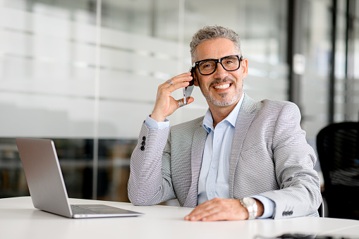 Headshot of handsome charismatic mature businessman talking on smartphone sitting at the office desk, middle-aged 50s man in formal wear having phone conversation with customers or business partners