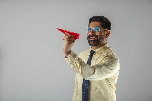 Mid adult smiling businessman dressed in formalwear flying paper airplane while standing against white background