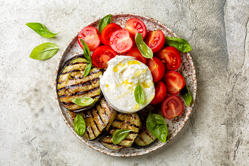 Plate with Burrata or mozzarella cheese and vegetables, grilled eggplant, tomatoes, basils, grissini and olive oil. Italian food, snack.