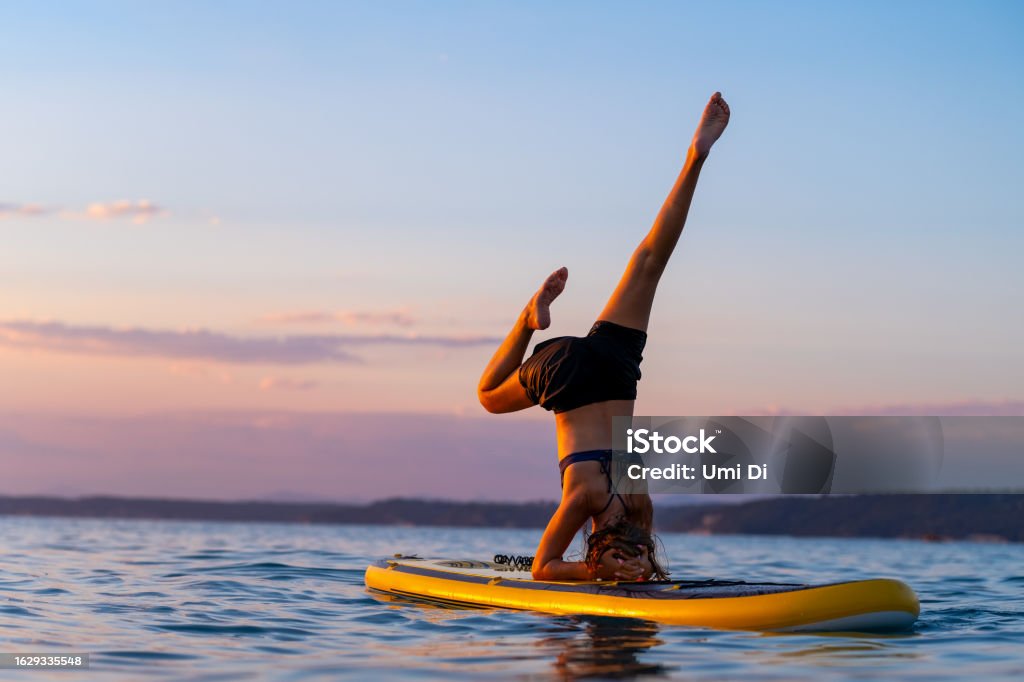 Perfect summer beach holidays, SUP yoga on a calm water of Aegean Sea Teenage girl doing headstand and stretching on a puddle board at sunset Paddleboard Stock Photo