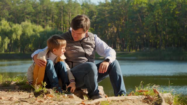 Little boy sitting with father at the lake and looking on the sunset over forest. Family outdoors, happy parenting and childhood, autumn landscape, kids and parents relaxing outside