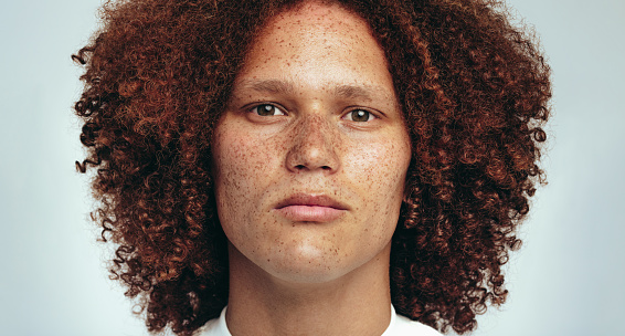 Portrait of a handsome young man with freckles and a curly afro looking at the camera in a studio, expressing confidence in his unique appearance. Youthful man embracing his natural beauty.