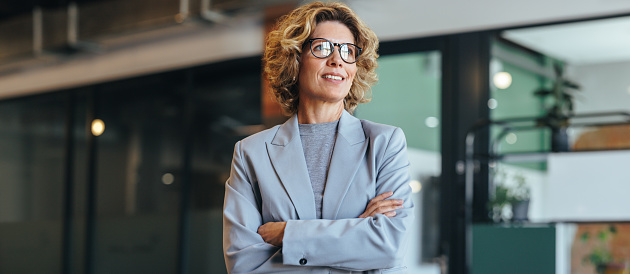 Thoughtful business woman standing with crossed arms. Woman in her 40's working in an office. Professional woman wearing a suit.
