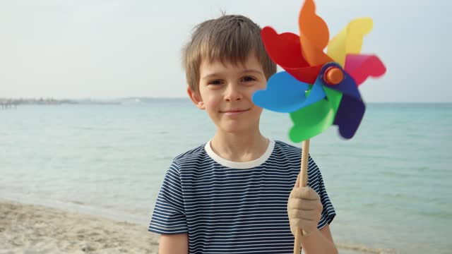 Portrait of happy smiling boy standing on sandy sea beach and holding colorful pinwheel toy. Concept of child happiness during summer vacation or weekend