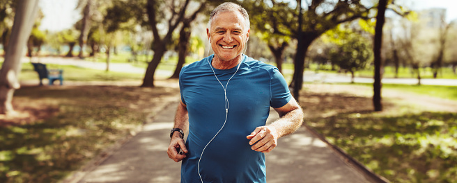 Senior man running in a park with a smile on his face and earphones on. Elderly man embracing physical fitness, caring for his health and wellbeing and longevity in his golden years.