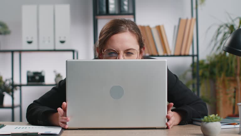 Confident young business woman hiding behind laptop computer, looking at camera, spying, peeping