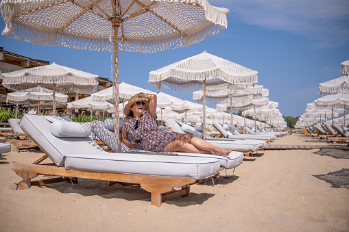 A smiling young woman wearing large sunglasses and a straw hat with an unraveled brim lying on a sun lounger against the background of many sun loungers and wicker beach umbrellas arranged in rows, a beach, and a blue sky with small white clouds. The wicker beach umbrella casts a patterned shadow on her body.