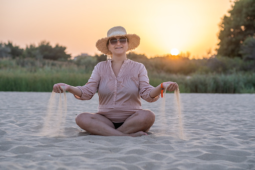 Young cheerful woman on a deserted beach at sunset. She is standing in the lotus position, she has taken sand in both hands and is letting it run through her fingers.