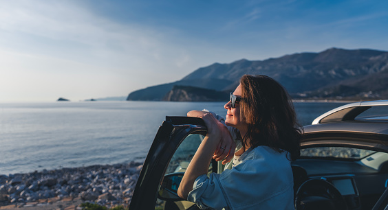 Young happy woman traveler enjoying the sunset at the sea while standing next to the car. Summer holidays and travel concept