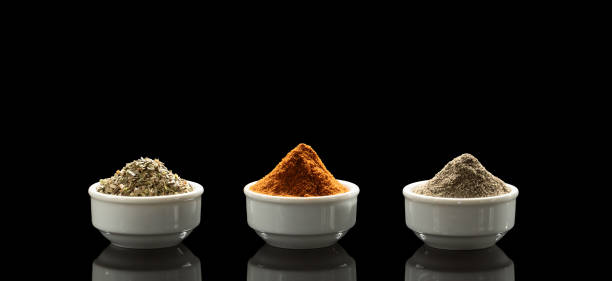 An overhead view of four stainless steel spoons arranged on a white background, each holding a different type of vibrant spice. The collection includes whole cubeb peppers (tailed pepper), cumin seeds, bright yellow turmeric powder, and rich red chili powder. This image represents a palette of flavors essential to many world cuisines, particularly Indian and Asian cooking.